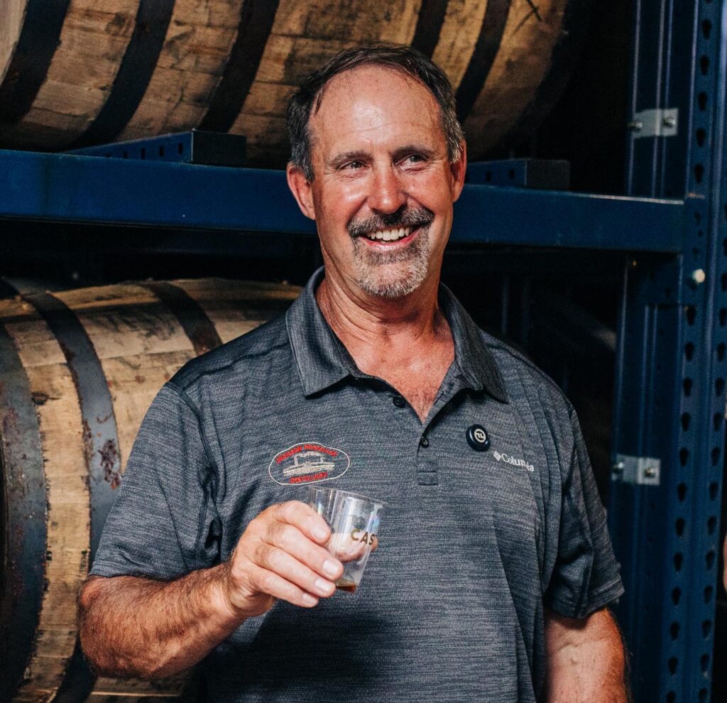 Craig Beam from Jackson Purchase Distillery holding a cup of bourbon with bourbon barrel shelves behind him